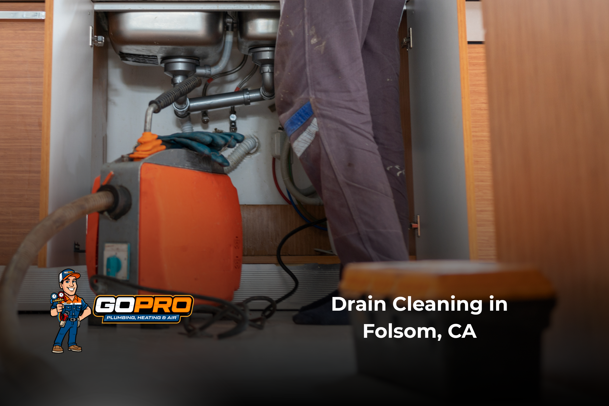 A plumber operates an orange drain-cleaning machine beneath a kitchen sink, with pipes and hoses visible; caption reads “Drain Cleaning in Folsom, CA.”